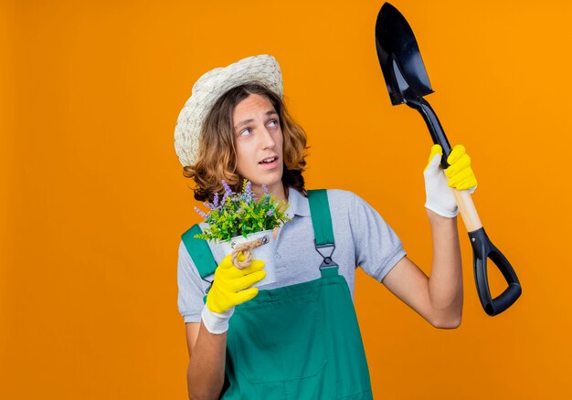 Young gardener man in rubber gloves wearing jumpsuit and hat holding shovel