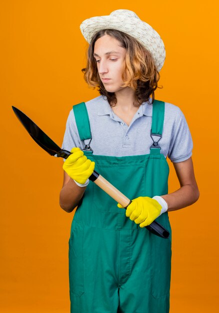 Young gardener man in rubber gloves wearing jumpsuit and hat holding shovel