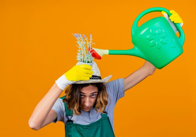 Young gardener man in rubber gloves wearing jumpsuit and hat holding watering can watering plant on his head standing over orange background