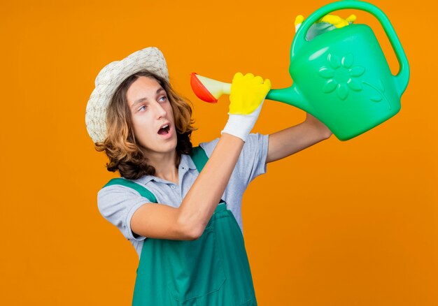 Young gardener man in rubber gloves wearing jumpsuit and hat holding watering can