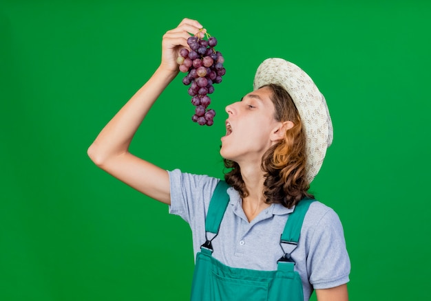 Бесплатное фото Молодой садовник в комбинезоне и шляпе держит гроздь винограда, открывая рот