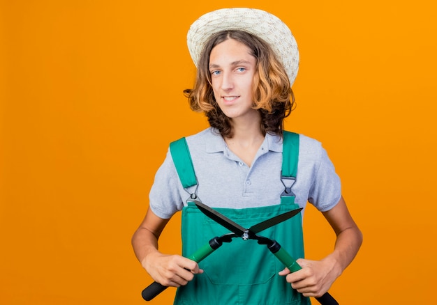 Бесплатное фото Молодой садовник в комбинезоне и шляпе с улыбкой держит ножницы для живой изгороди