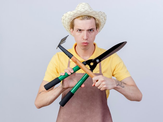 Бесплатное фото Молодой садовник в комбинезоне и шляпе держит мини-грабли и ножницы для живой изгороди, скрестив руки с серьезным лицом