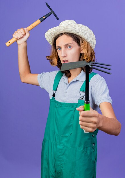 Бесплатное фото Молодой садовник в комбинезоне и шляпе с мини-граблями и мотыгой