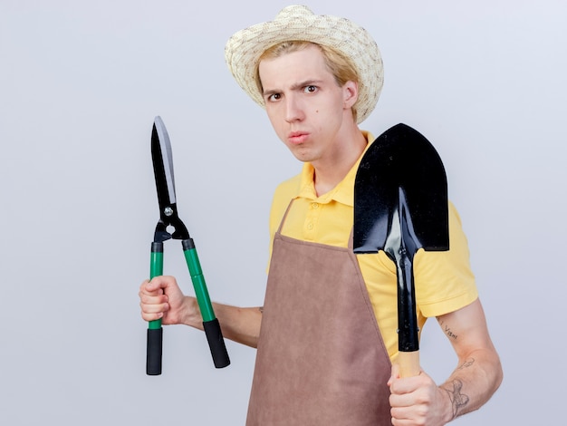 Бесплатное фото Молодой садовник в комбинезоне и шляпе держит лопату и ножницы для живой изгороди с серьезным лицом