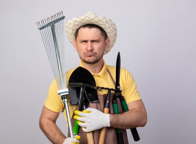 無料写真 深刻な眉をひそめている顔で園芸機器を保持している作業用手袋でジャンプスーツと帽子を身に着けている若い庭師の男
