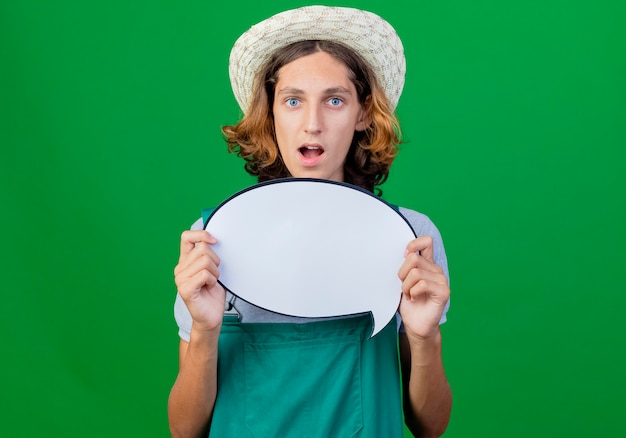 Young gardener man wearing jumpsuit and hat holding blank speech bubble sign