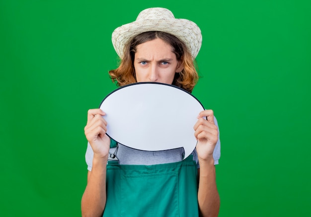 Young gardener man wearing jumpsuit and hat holding blank speech bubble sign