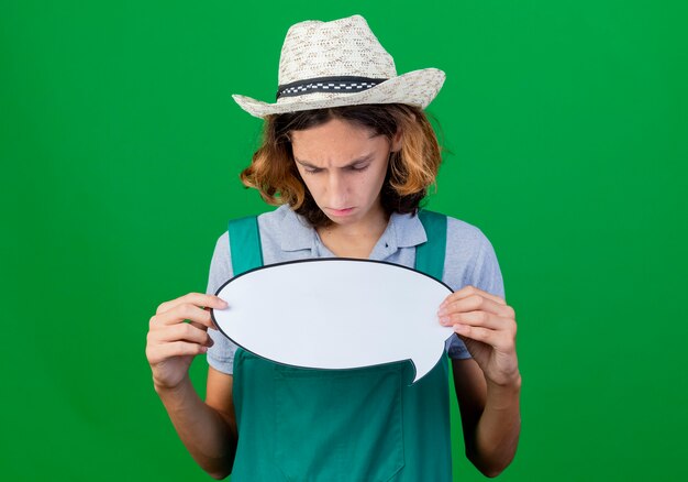 young gardener man wearing jumpsuit and hat holding blank speech bubble sign