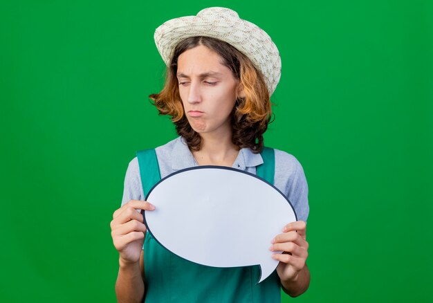 Young gardener man wearing jumpsuit and hat holding blank speech bubble sign