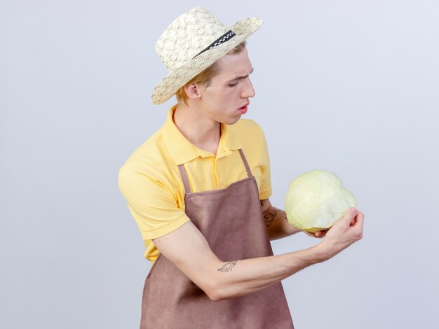 Young gardener man wearing jumpsuit and hat holding cabbage looking at it surprised 