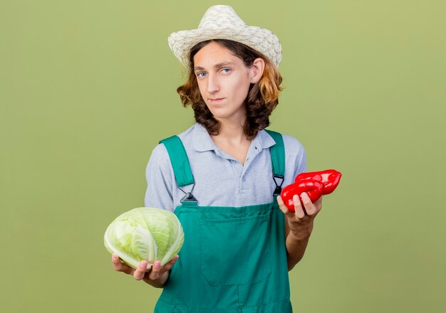 Young gardener man wearing jumpsuit and hat holding cabbage and red bell peppers