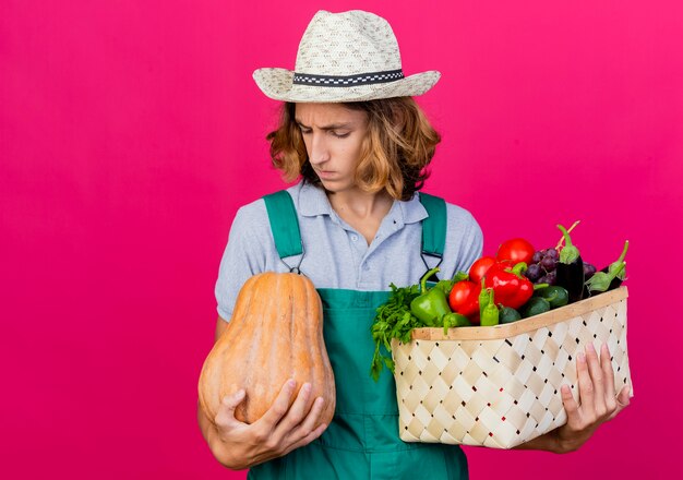 Young gardener man wearing jumpsuit and hat holding crate full of fresh vegetables