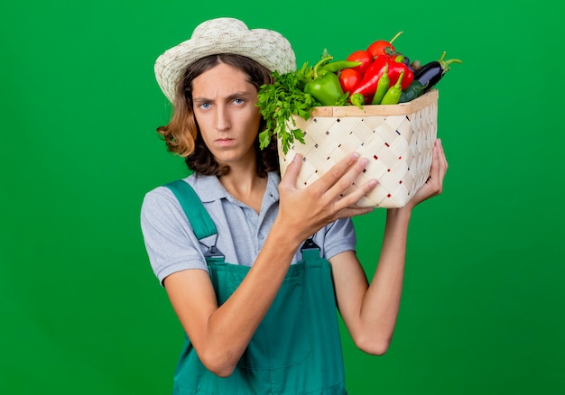 Young gardener man wearing jumpsuit and hat holding crate full of fresh vegetables