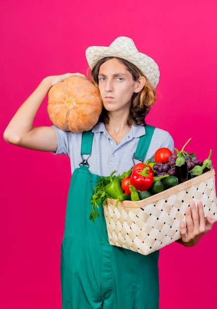 Young gardener man wearing jumpsuit and hat holding crate full of fresh vegetables