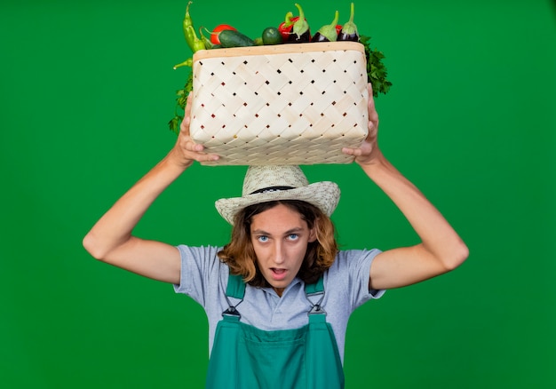 Free photo young gardener man wearing jumpsuit and hat holding crate full of fresh vegetables