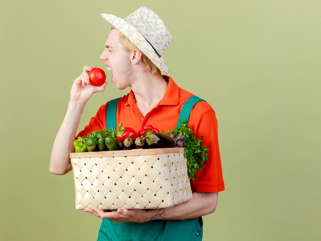 Young gardener man wearing jumpsuit and hat holding crate full of vegetables biting fresh tomato standing over light background