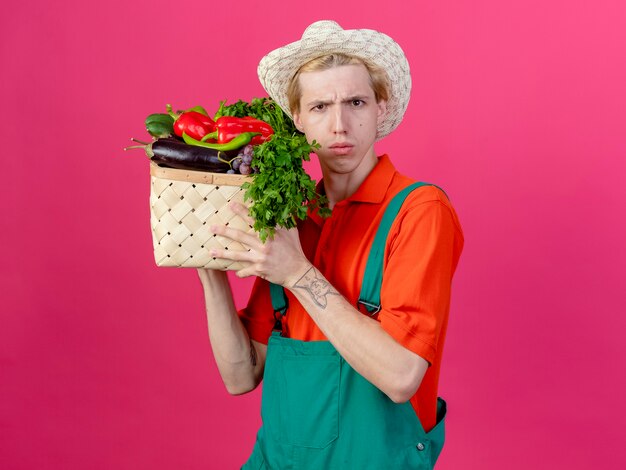 Молодой садовник в комбинезоне и шляпе держит ящик, полный овощей