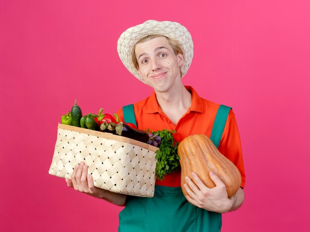 野菜でいっぱいの木枠を保持しているジャンプスーツと帽子を身に着けている若い庭師の男