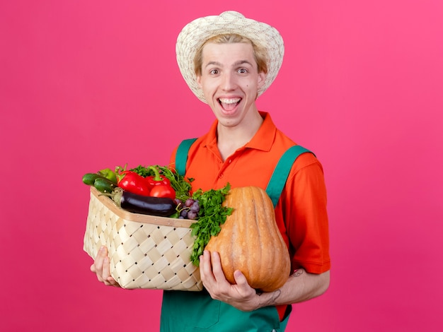 Free photo young gardener man wearing jumpsuit and hat holding crate full of vegetables