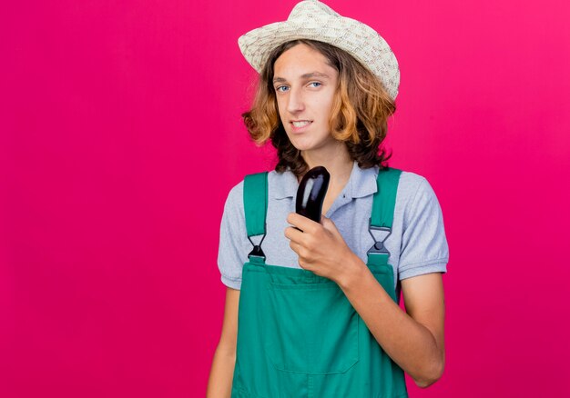 Young gardener man wearing jumpsuit and hat holding fresh eggplant