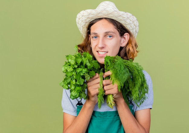 Young gardener man wearing jumpsuit and hat holding fresh herbs