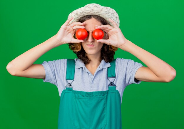 目を覆う新鮮なトマトを保持しているジャンプスーツと帽子を身に着けている若い庭師の男