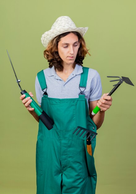 Young gardener man wearing jumpsuit and hat holding gardening equipment