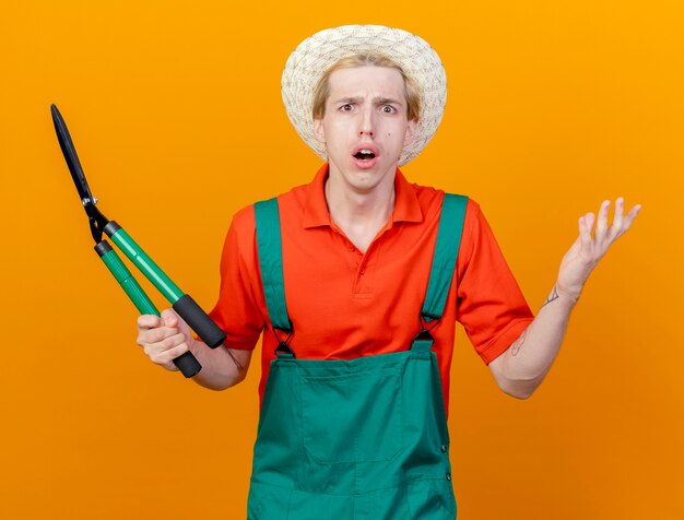 Young gardener man wearing jumpsuit and hat holding hedge clippers being confused