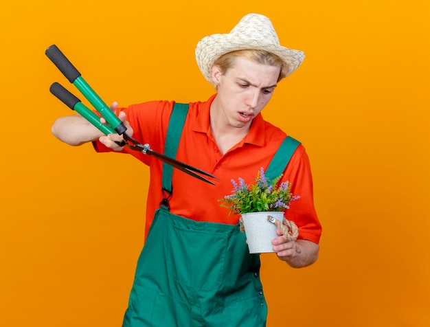 Free photo young gardener man wearing jumpsuit and hat holding hedge clippers and potted plant