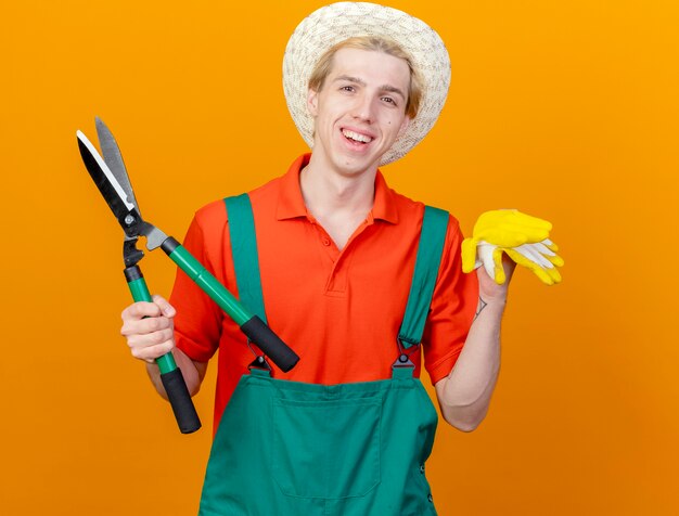 Young gardener man wearing jumpsuit and hat holding hedge clippers and rubber gloves looking at camera smiling cheerfully with happy face standing over orange background