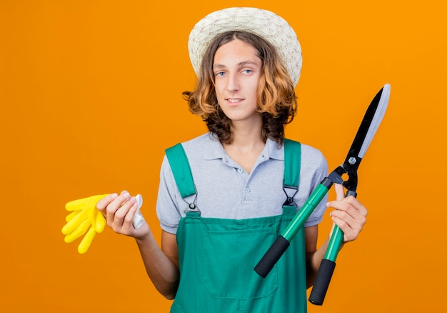 Giovane giardiniere uomo che indossa tuta e cappello tenendo tagliasiepi e guanti di gomma con il sorriso