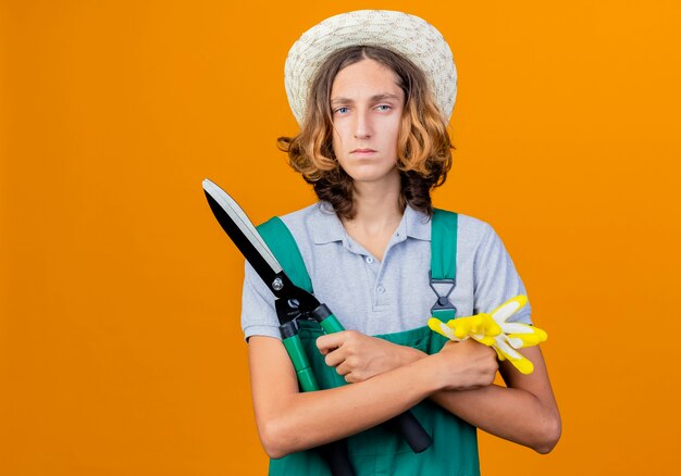 Young gardener man wearing jumpsuit and hat holding hedge clippers and rubber gloves