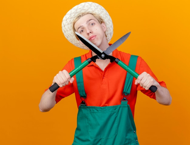 Young gardener man wearing jumpsuit and hat holding hedge clippers with serious face