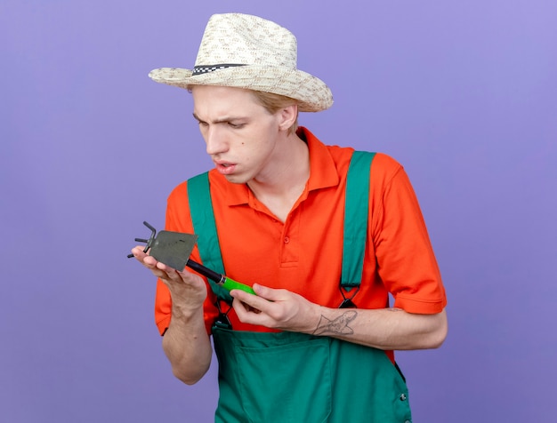 Young gardener man wearing jumpsuit and hat holding mattock looking at it intrigued standing over purple background