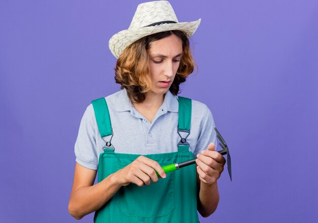 Young gardener man wearing jumpsuit and hat holding mattock looking at it serious