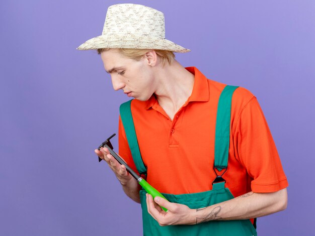 Young gardener man wearing jumpsuit and hat holding mattock looking at it with smile