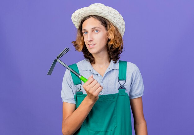 Free photo young gardener man wearing jumpsuit and hat holding mini rake looking at camera