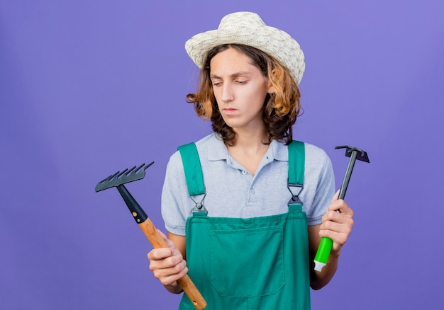 Young gardener man wearing jumpsuit and hat holding mini rake and mattock