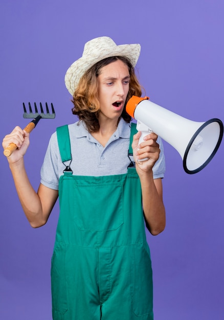 Young gardener man wearing jumpsuit and hat holding mini rake and shouting