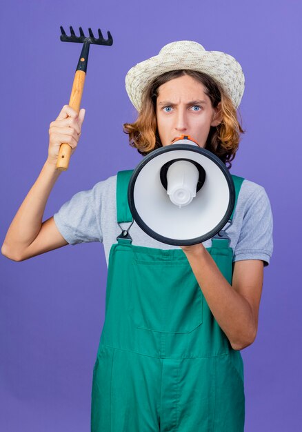 Young gardener man wearing jumpsuit and hat holding mini rake shouting