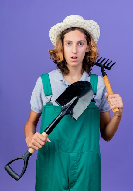 Молодой садовник в комбинезоне и шляпе держит мини-грабли и лопату с серьезным лицом