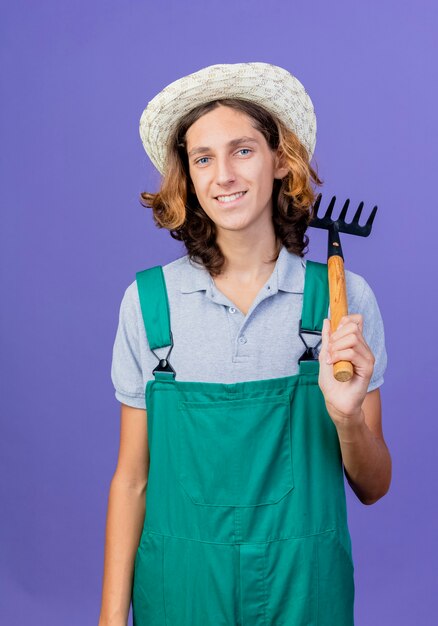 Young gardener man wearing jumpsuit and hat holding mini rake smiling cheerfully