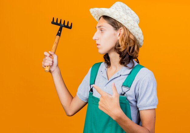 Young gardener man wearing jumpsuit and hat holding mini rake