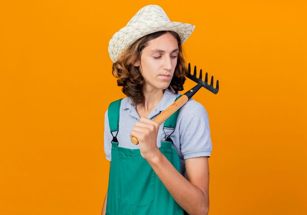 Young gardener man wearing jumpsuit and hat holding mini rake