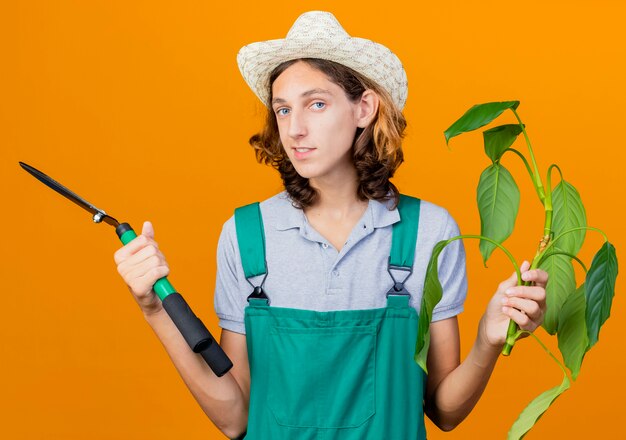 Young gardener man wearing jumpsuit and hat holding plant and hedge clippers