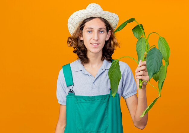 Young gardener man wearing jumpsuit and hat holding plant