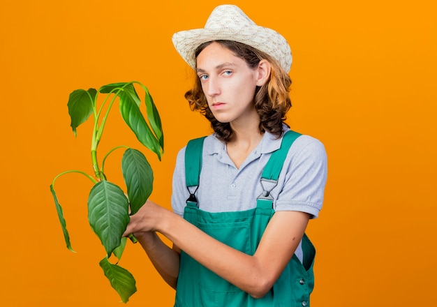 Молодой садовник в комбинезоне и шляпе держит растение