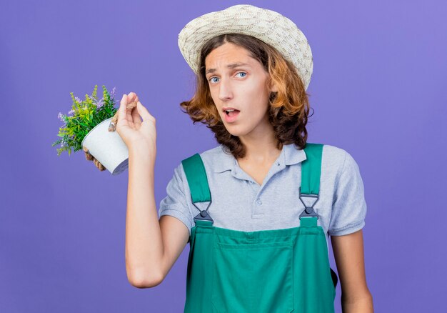 Young gardener man wearing jumpsuit and hat holding potted plant being confused