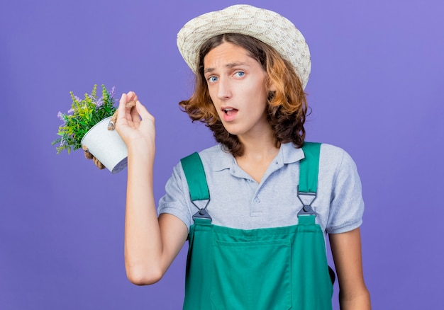 混乱している鉢植えの植物を保持しているジャンプスーツと帽子を身に着けている若い庭師の男
